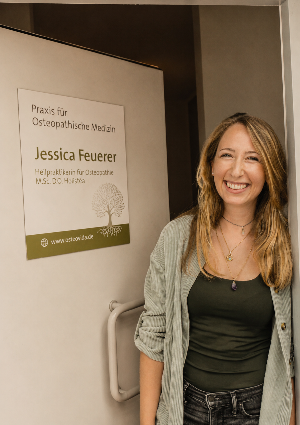 Jessica Feuerer begrüßt in der geöffneten Tür der Osteopathiepraxis OsteoVida